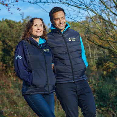 A dark haired gentleman wears a navy padded body warmer with the Guide Dogs logo in yellow on the upper left corner and a light blue hoodie. A lady next to him is wearing the same body warmer.