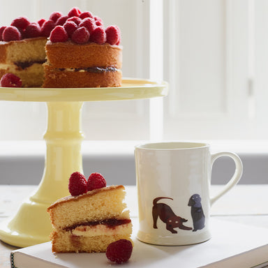A white ceramic mug sits on a book next to a piece of cake with jam and cream. The mug has an illustration of two days playing, one Chocolate Labrador and Black Labrador. The rest of the cake is on a cream cake stand behind the piece of cake.