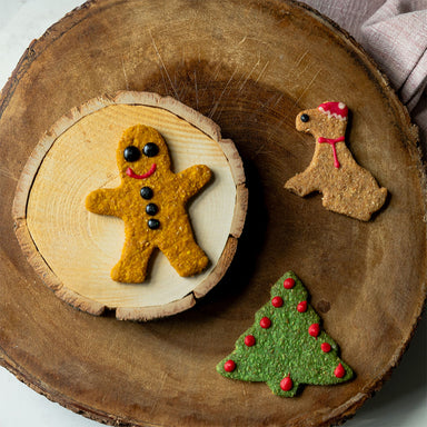 Three dog biscuits in festive shapes, a Christmas tree, gingerbread man and dog with a Santa hat. They are placed on a wooden surface.