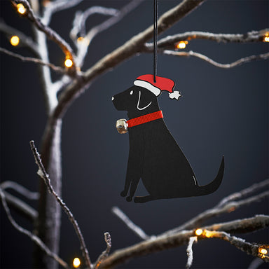 A wooden Black Labrador decoration with red Santa hat and red ribbon collar and bell. The decoration is hanging on a tree with lights.