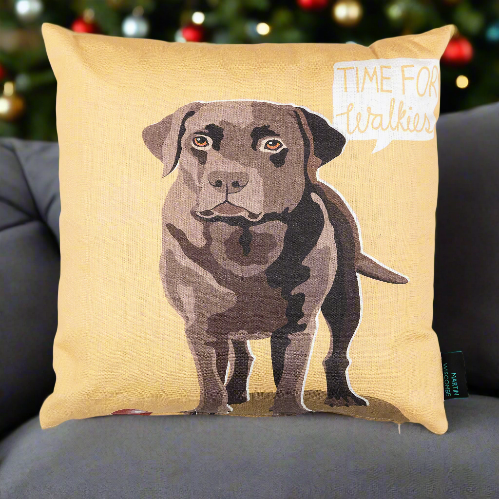 A cushion with illustrated chocolate Labrador on a yellow background, with the words 'Time for Walkies!' in the top corner. The cushion is on a sofa with a Christmas tree in the background.