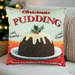 A cushion with illustrated Christmas pudding design, pale green background. The cushion is on a sofa with a Christmas tree in the background.