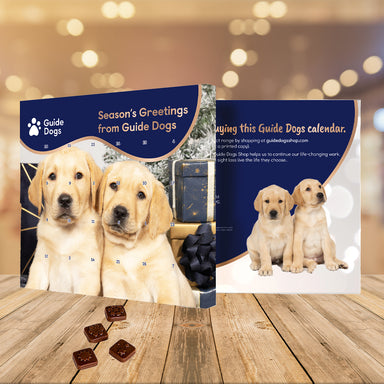 An advent calendar featuring two Yellow Labrador puppies. The calendar has 24 windows and square shaped chocolate are below as an example. The calendar rests of a wooden table with gold lights in the background.