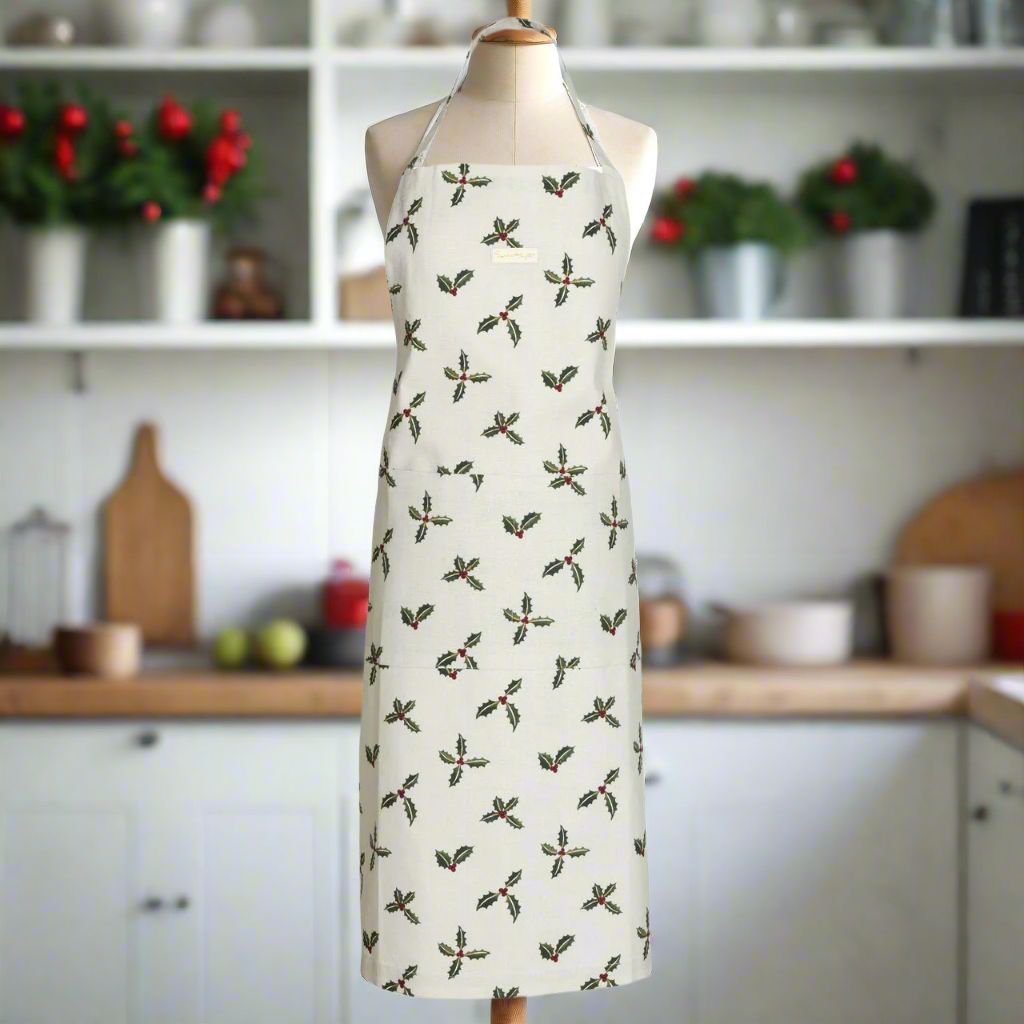 A white apron decorated with a repeating pattern of holly with berries. The apron is on a mannequin in a kitchen.