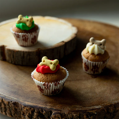 A photograph of three mini iced cupcakes for dogs. One has green icing, one has white icing and one has red icing. They have been placed on a wooden surface.