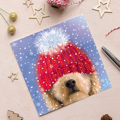 A Christmas card featuring an illustration of a golden dog with a red bobble hat pulled down over their eyes. The card is on a blue background with white snowflakes.