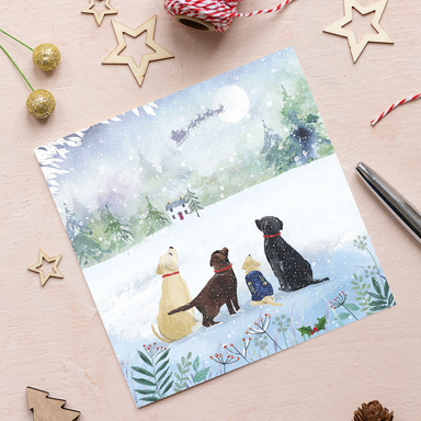 A Christmas card featuring four Labradors sitting in the snow watching Father Christmas's sleigh cross the moon.