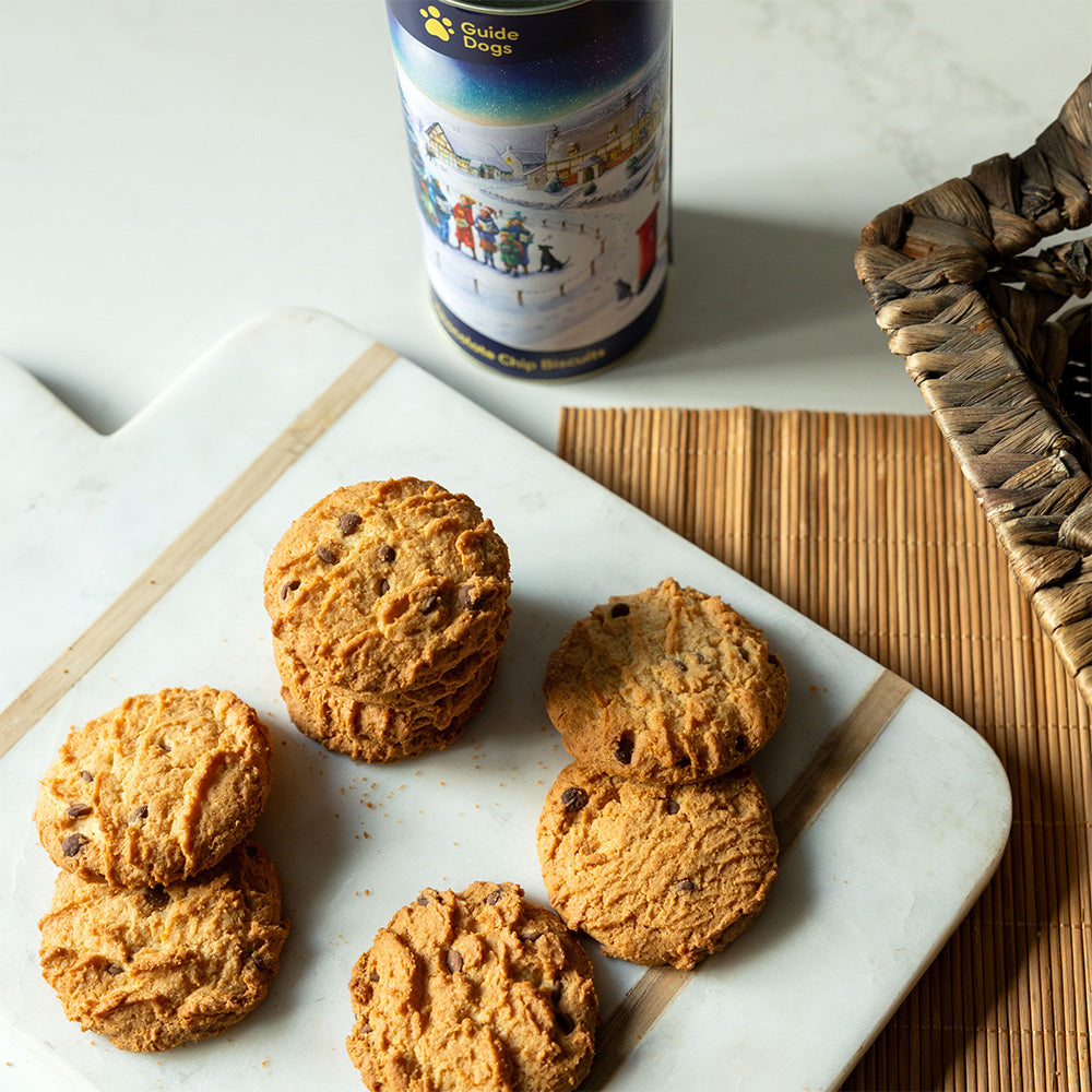 A photograph of chocolate chip cookies in front of a cylinder biscuit tin featuring a snowy village illustration.