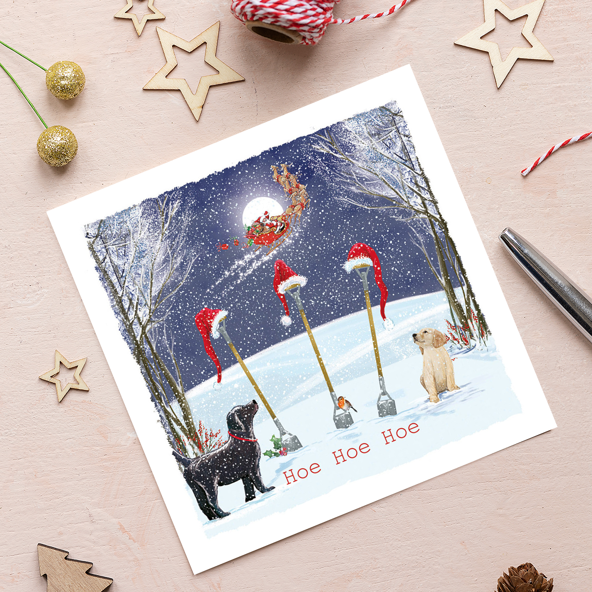 A Christmas card featuring an illustration of three garden hoe tools in snow with Santa hats on top, with the phrase ‘Hoe Hoe Hoe’ appearing below. The spelling of Hoe is to match the garden tool, in jest of the common Christmas phrase ‘Ho Ho Ho’. The card also features a Black Labrador stood and Yellow Labrador sat by the tools watching Father Christmas and his reindeer in the night sky. 