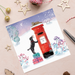 An illustration of a Black Labrador puppy with a letter in its mouth, trying to reach a red post box. The puppy is in the snow, stood on some presents to give extra height. A cat is laying on top of the post box.