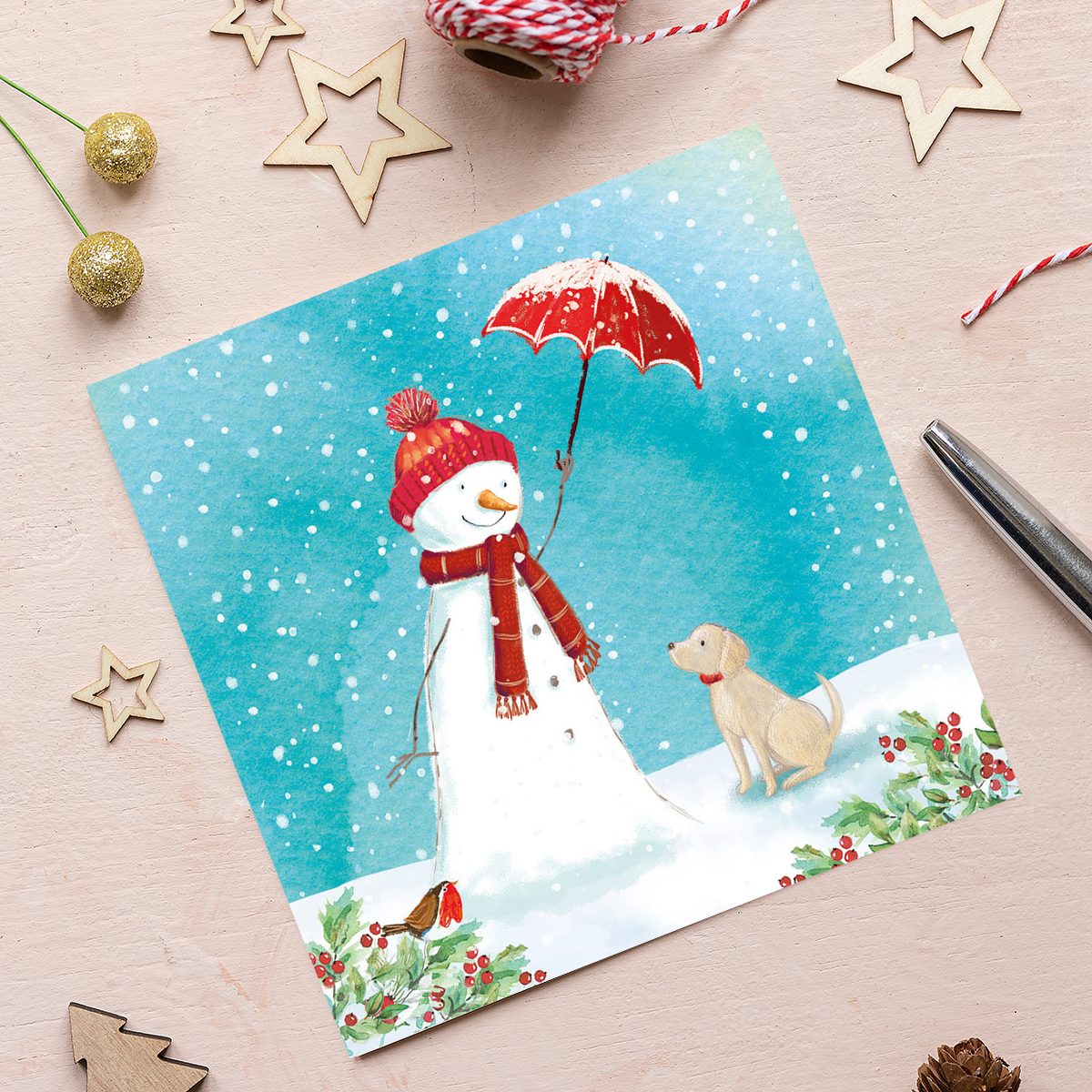 A Christmas card featuring an illustration of a snowman wearing a red woolly hat and scarf, holding a red umbrella to protect a Yellow Labrador puppy from the snow. There is holly and berries in the bottom corners with a robin watching the scene.