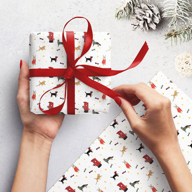 A woman's hands holding a wrapped present with another piece next to her. The wrap is white with illustrations of festive dogs and post boxes.