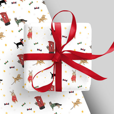 A piece of wrapping paper appears underneath an already wrapped present with red ribbon. The wrap is white with illustrations of festive dogs and post boxes. 