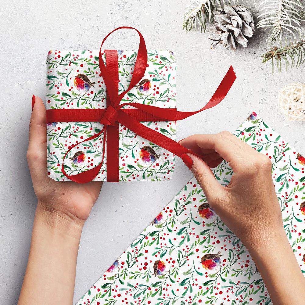 A woman's hands holding a wrapped present with another piece next to her. The wrap is white with illustrations of water colour robins amongst leaves and berries.