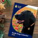 Front cover for the Guide Dogs 2025 Wall Calendar featuring a Black Labrador sitting in a field of poppies, which is on a wooden Christmassy background.