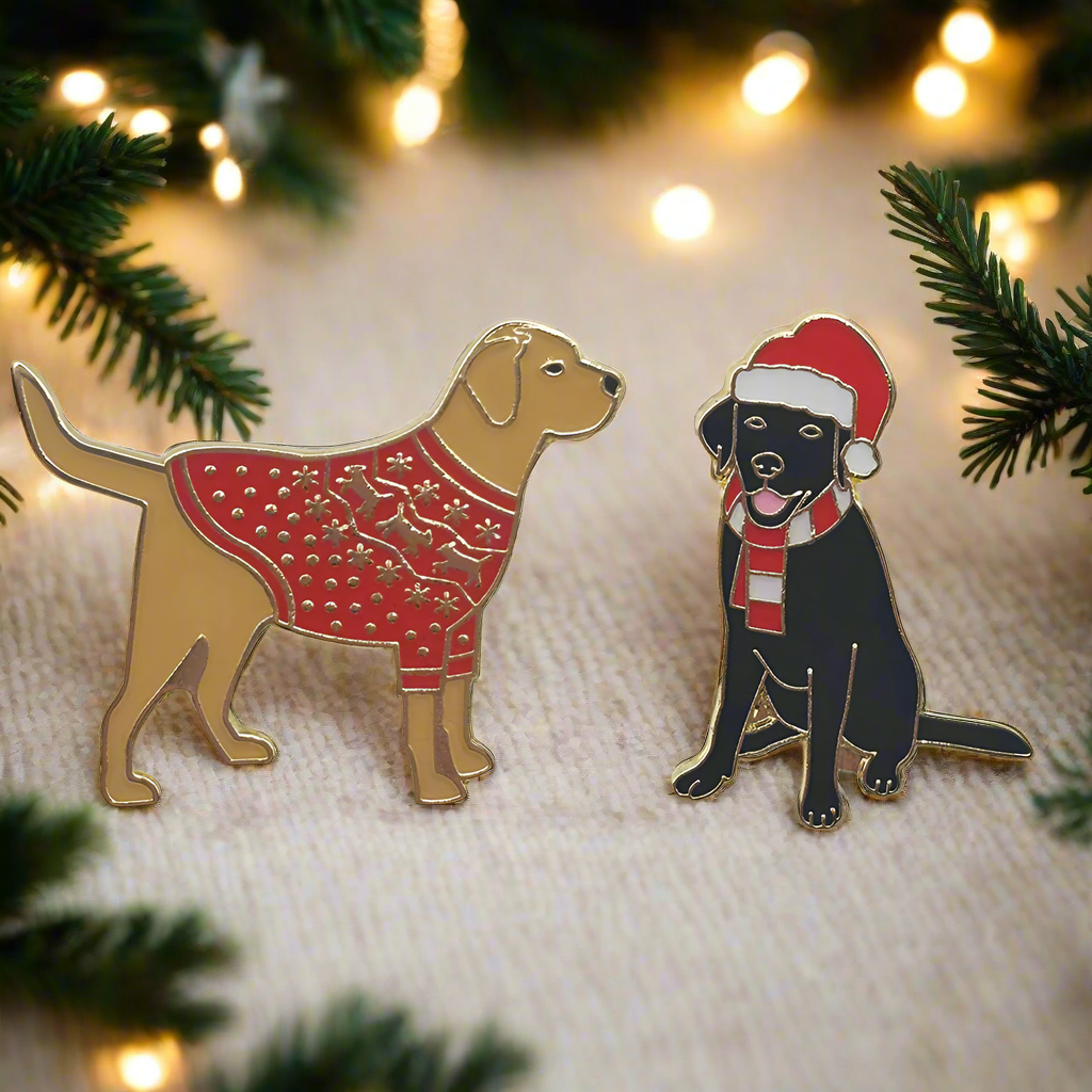 Two pin badges, one featuring a golden Labrador wearing a red Christmas jumper, the other featuring a black Labrador wearing a Santa hat and red and white stripe scarf. The background features pine branches and Christmas lights.