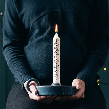 A white advent candle with numbers 1 to 24 down the front and small illustrations of different dog breeds. The candle is in a ceramic holder, being held by a person sitting down.