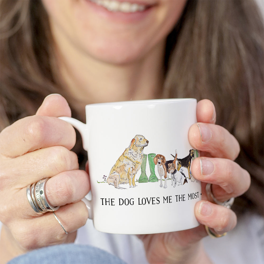 A photograph of a woman holding a white mug featuring various dog breeds with the text 'The dog loves me the most' underneath.