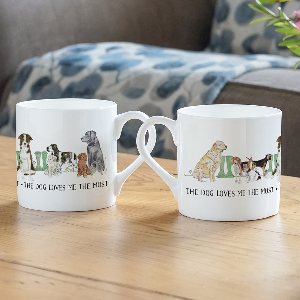 A photograph of two white mugs featuring various dog breeds with the text 'The dog loves me the most' underneath. The mugs are on a wooden table, showing off different sides of the mug.