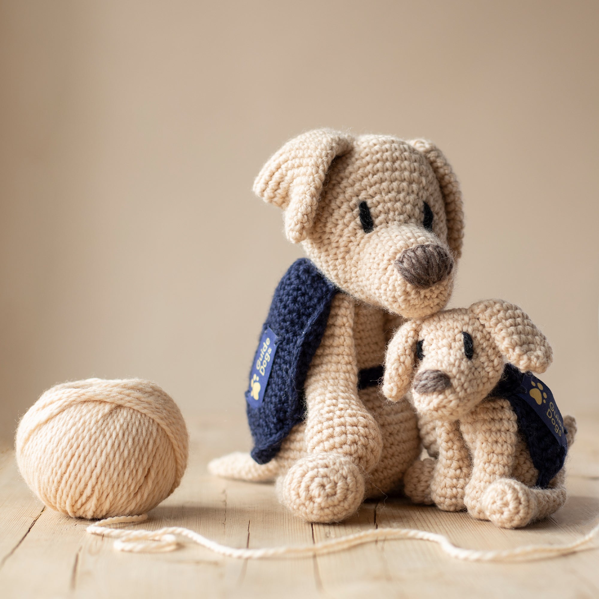 Two crocheted Yellow Labradors with Guide Dogs jackets, one large and one mini. Next to them is ball of yarn.