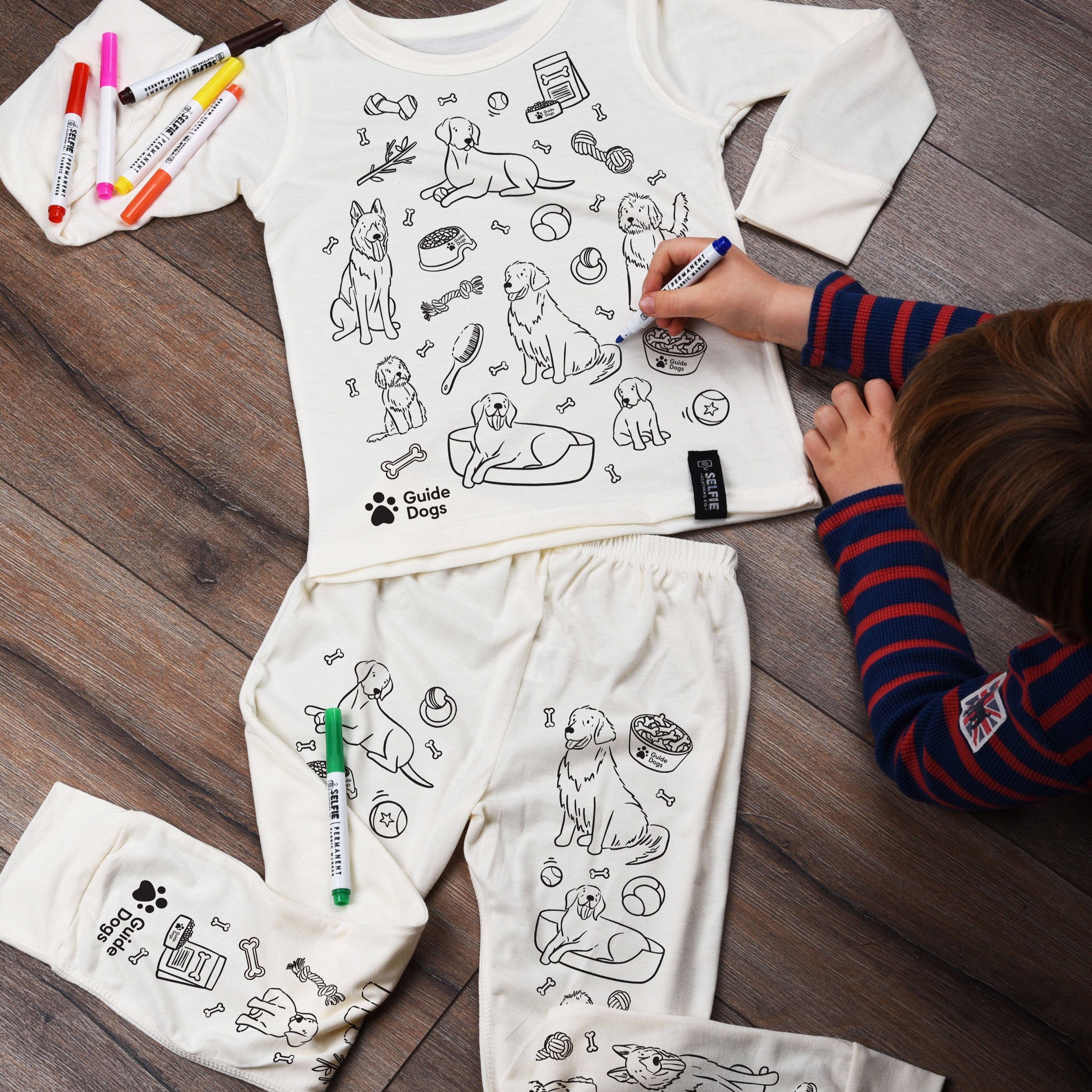 A photograph of a child colouring in Guide Dogs pyjamas. There is a long sleeved top and trousers featuring illustrations of dogs and other dog related items like toys, bones and bowls. There are seven colourful pens to choose from.