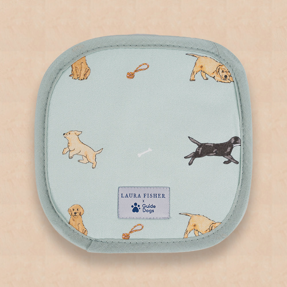 A pale blue pot grab with pattern of playing Labrador puppies and Guide Dogs label logo on a peach background.
