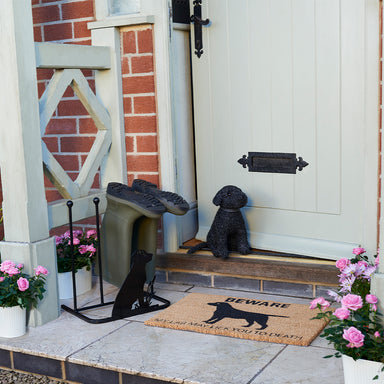 A photograph of the front of a house. The front door has a Black Labrador door stop to keep it ajar. There is a coir doormat in front the door. There is also a Black Labrador themed welly rack with a pair of khaki welly boots on.