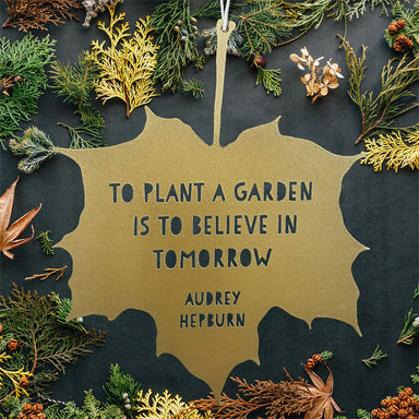 A gold hanging leaf with the quote 'to plant a garden is to believe in tomorrow'. Audrey Hepburn's name appears below. The background features autumnal leaves. 