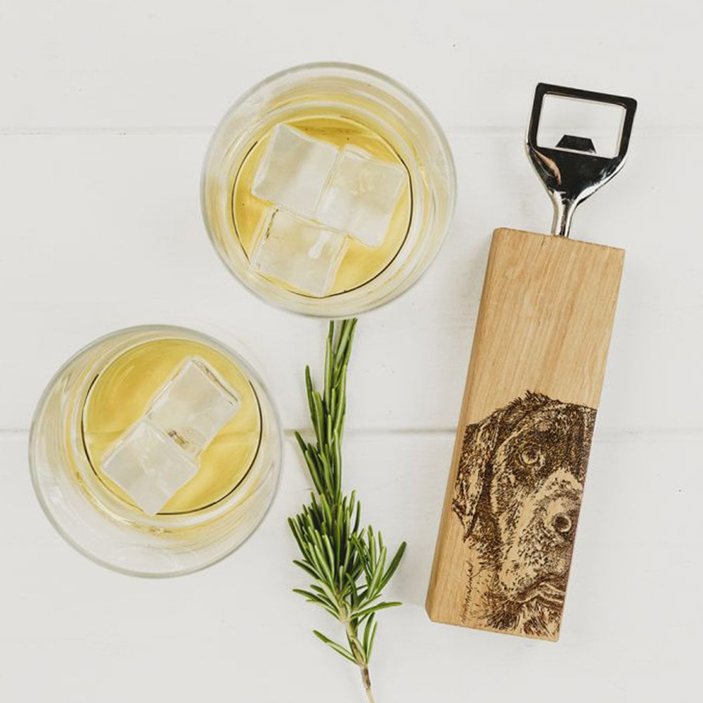 A metal bottle opener with wooden base. The base has an engraved illustration of a Labrador. There are two drinks served with ice next to the bottle opener.