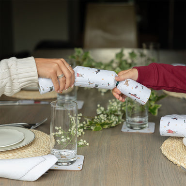 A photograph of two people pulling a cracker featuring an illustrated festive dogs design over a Christmas table.