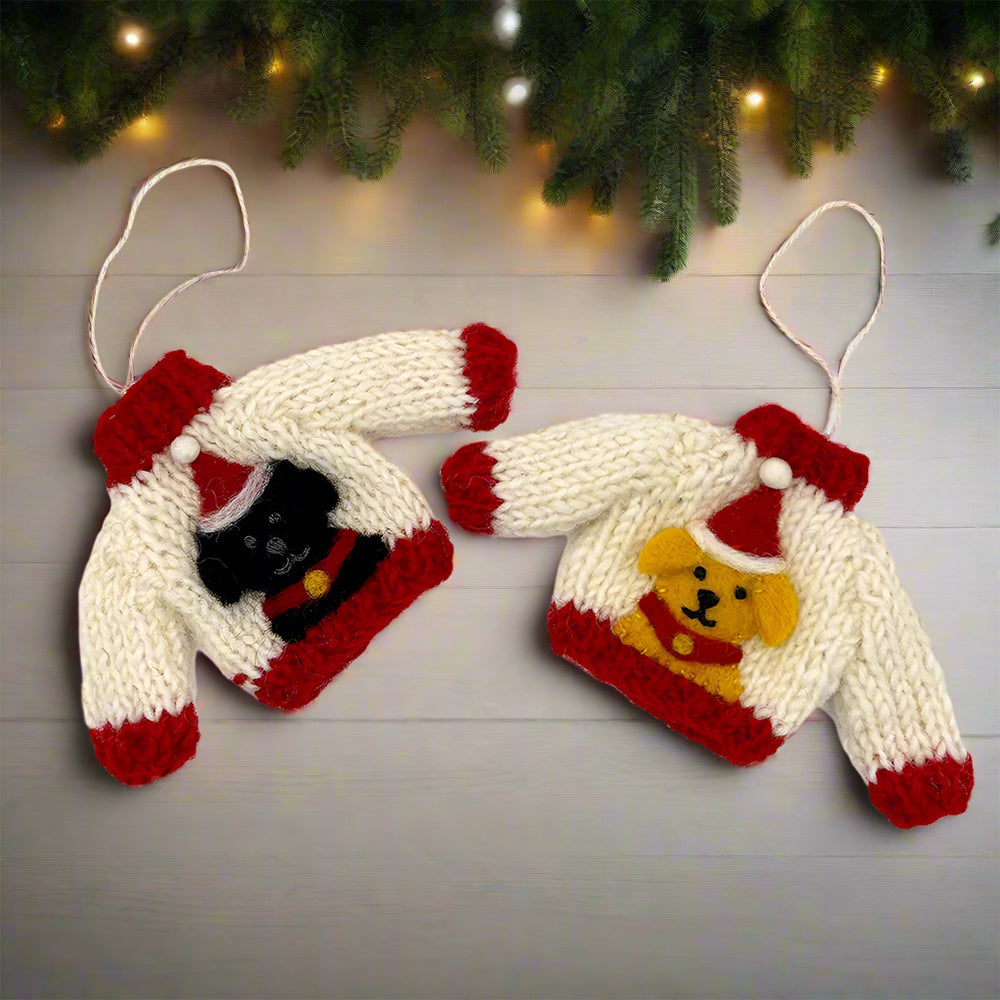 Two knitted Christmas jumper decorations, one featuring a black Labrador, the other a golden Labrador. Both jumpers are white with red edges and the Labradors are wearing a Santa hat. They are on a wooden floor in front of pine branches and fairy lights.