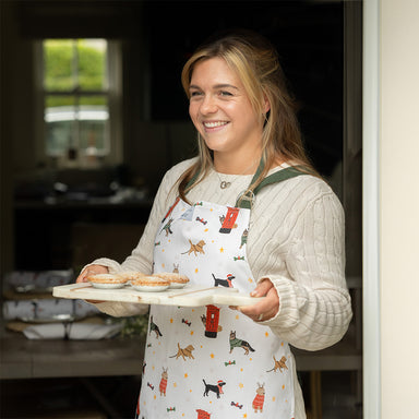 A woman wearing a white apron with dark green straps. The apron features a printed design with different dog breeds and post-boxes at Christmas. She is in the doorway holding a tray of mince pies.