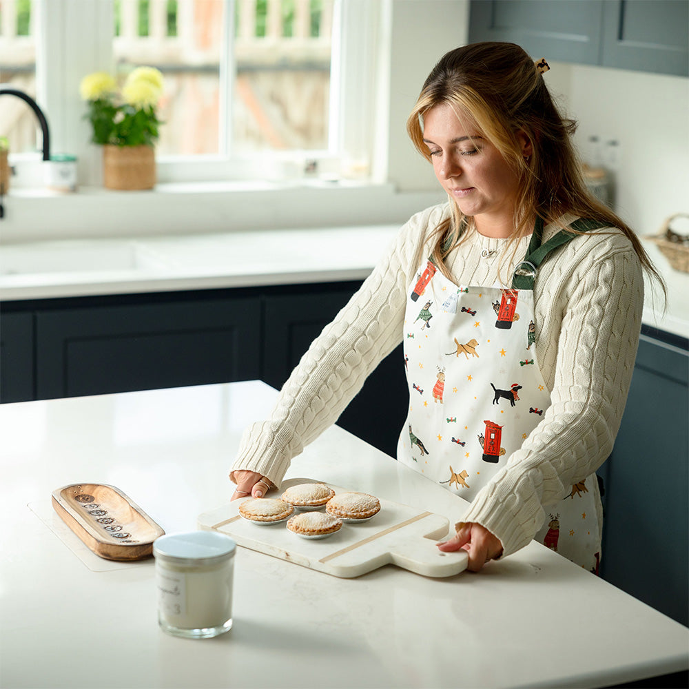 A woman wearing a white apron with dark green straps. The apron features a printed design with different dog breeds and post-boxes at Christmas. She is in the kitchen holding a tray of mince pies.