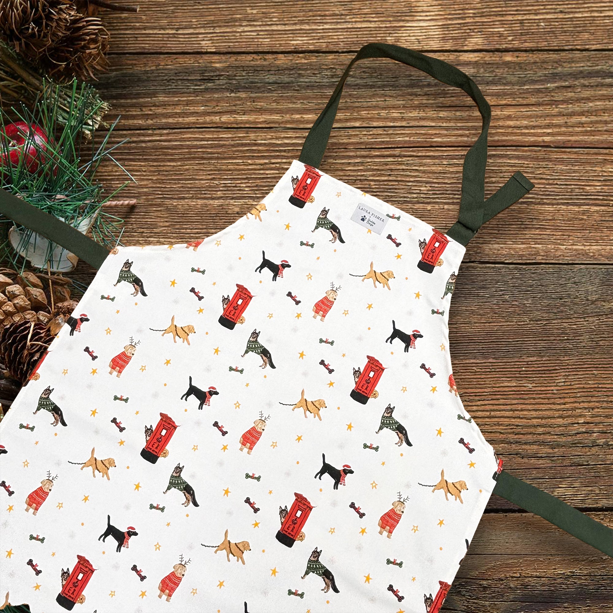 A white apron with dark green straps. The apron features a printed design with different dog breeds and post-boxes at Christmas.