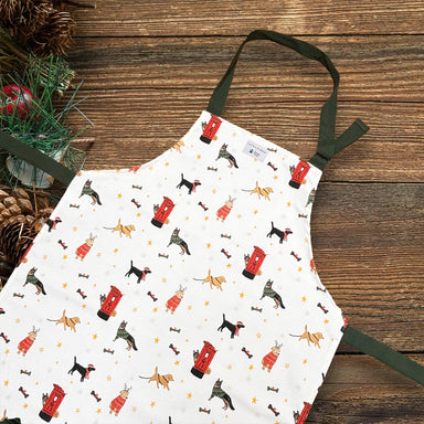 A white apron with dark green straps. The apron features a printed design with different dog breeds and postboxes at Christmas.