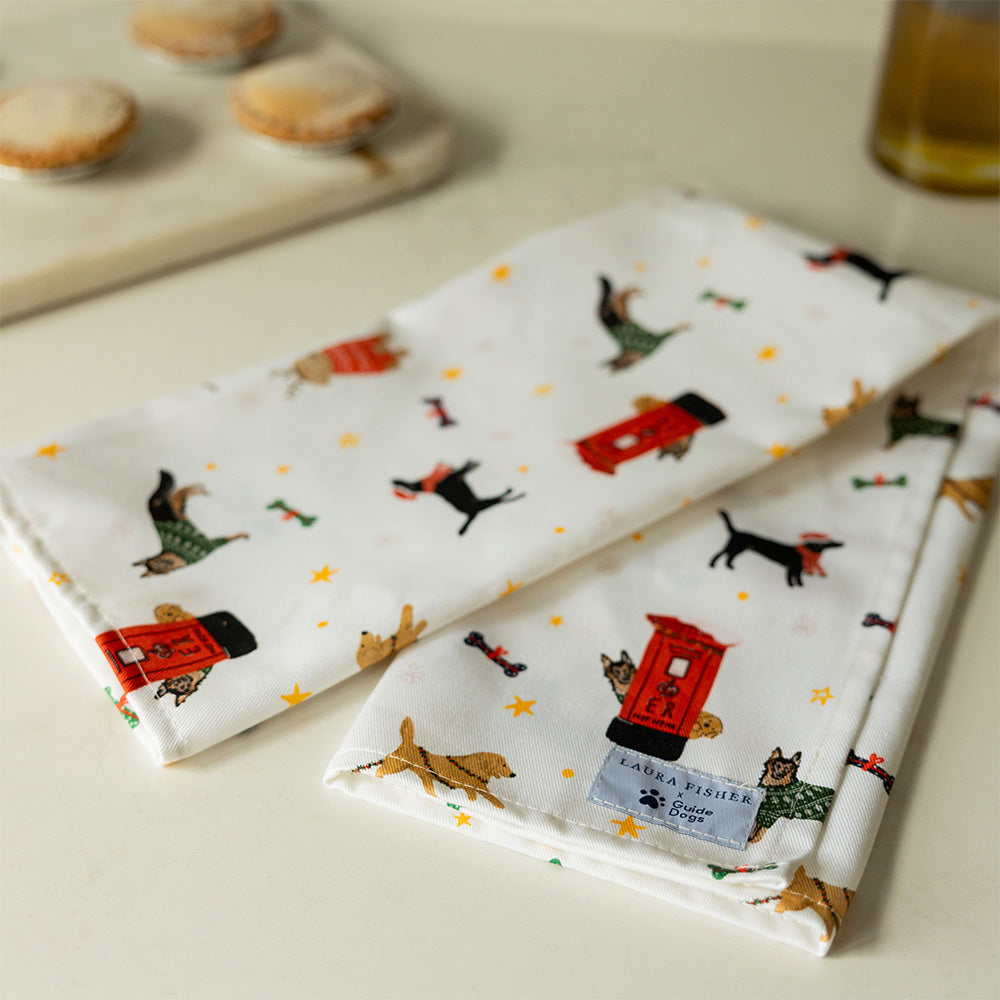 A folded white tea towel featuring a printed design with different dog breeds and post-boxes at Christmas. The tea towel is on a table with mince pies in the background.