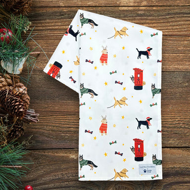 A folded white tea towel featuring a printed design with different dog breeds and post-boxes at Christmas. The tea towel is on a wooden table with pinecones on the side.