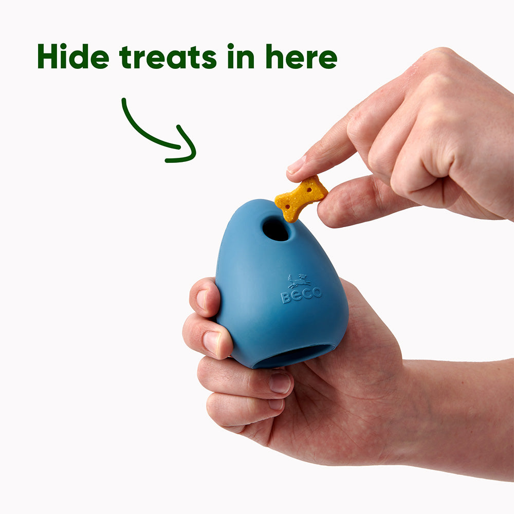 A person holding a blue rubber toy for dogs that can be filled with treats. The person is about to add a dog treat to it. There's an arrow pointing to the top hole with the words 'Hide treats in here'.