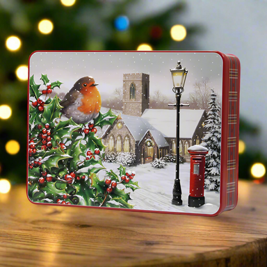 A photograph of a tin of biscuits featuring a robin and a church on a snowy evening. The tin is on a wooden table with a Christmas tree in the background.