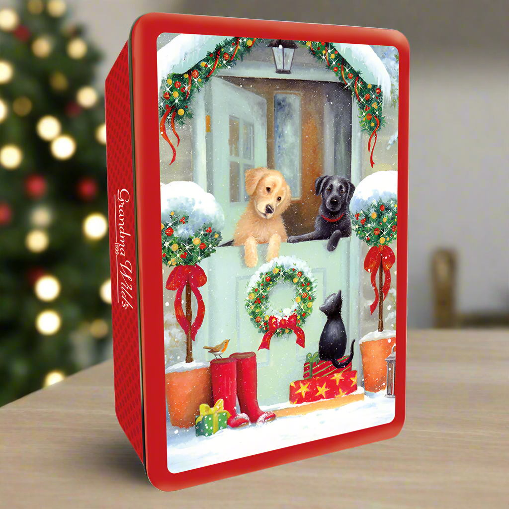 A photograph of a biscuit tin featuring two Labradors peeking over a gate at a festive scene including a black cat. The tin is on a wooden table with a Christmas tree in the background.