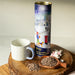 A photograph of a tall cylinder tin with a festive illustration on a wooden surface. There are hot chocolate flakes on a wooden spoon, mini marshmallows and a mug next to the cylinder.