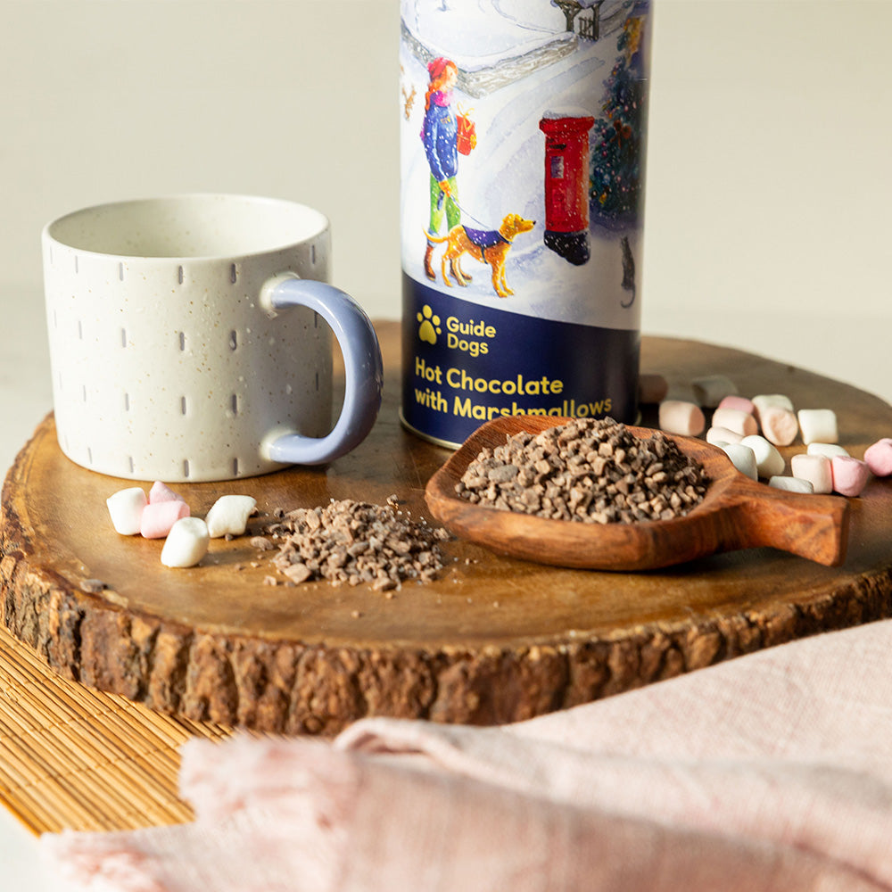 A close-up photograph of a cylinder tin with a festive illustration on a wooden surface. There are hot chocolate flakes on a wooden spoon, mini marshmallows and a mug next to the cylinder.