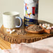 A close-up photograph of a cylinder tin with a festive illustration on a wooden surface. There are hot chocolate flakes on a wooden spoon, mini marshmallows and a mug next to the cylinder.