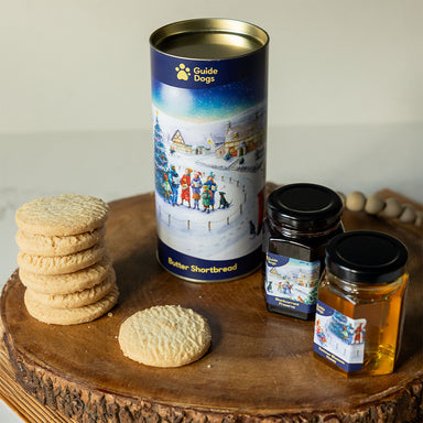 A photograph of the teatime treats gift set on a wooden table. The set is made up of a cylindrical tin of butter shortbread and jars of blackcurrant preserve and summer blossom honey. Pieces of round shortbread are next to the tin.