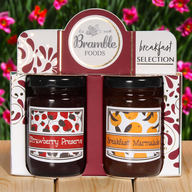 A breakfast selection featuring a jar of strawberry preserve and a jar of breakfast marmalade in red cardboard packaging. There are pink flowers in the background.