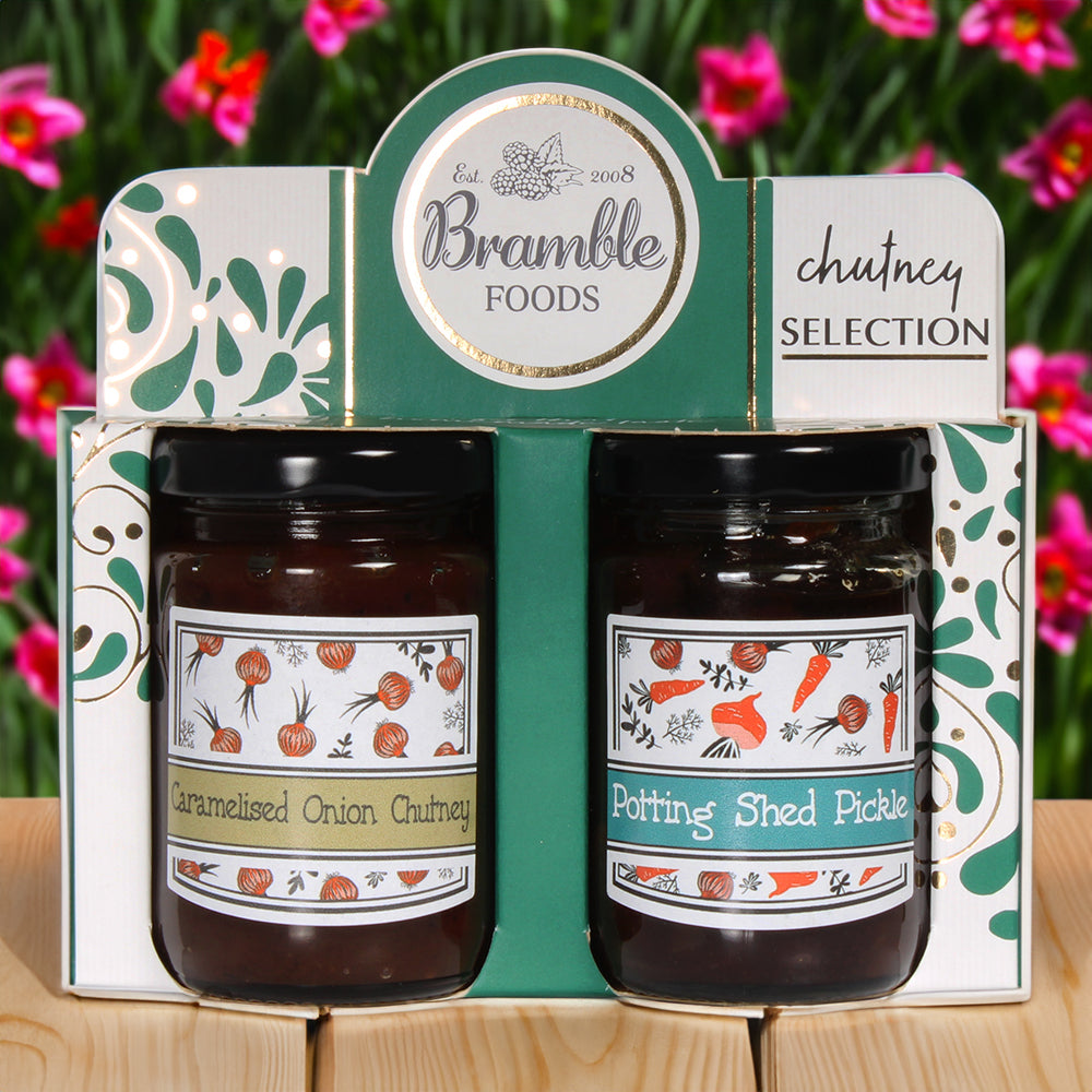 A chutney selection featuring a jar of caramelised onion chutney and a jar of potting shed pickle in green cardboard packaging. There are pink flowers in the background.