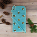 A teal glasses case with a repeated bees pattern. The case is on a wooden table with pine cones.