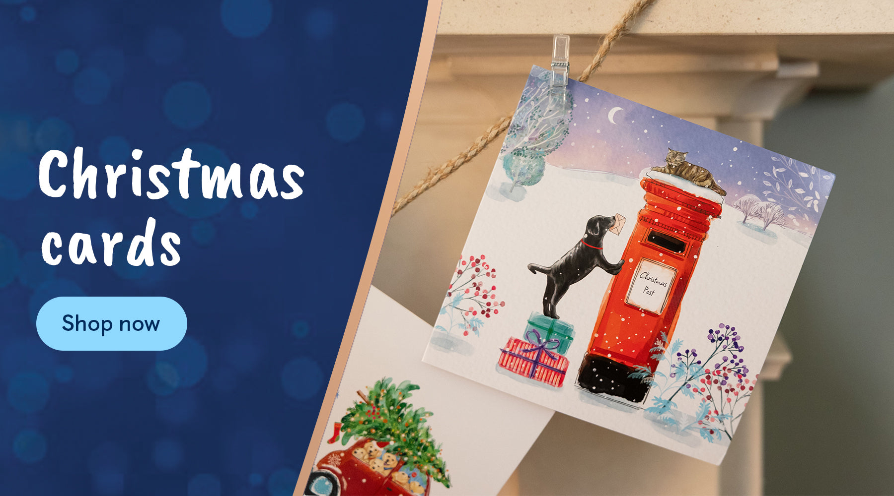 A photograph of a Christmas card featuring an illustration of a Black Labrador puppy with a letter in its mouth, trying to reach a red post box is clipped to string on a mantelpiece. There is an ask to shop via the Christmas cards category.