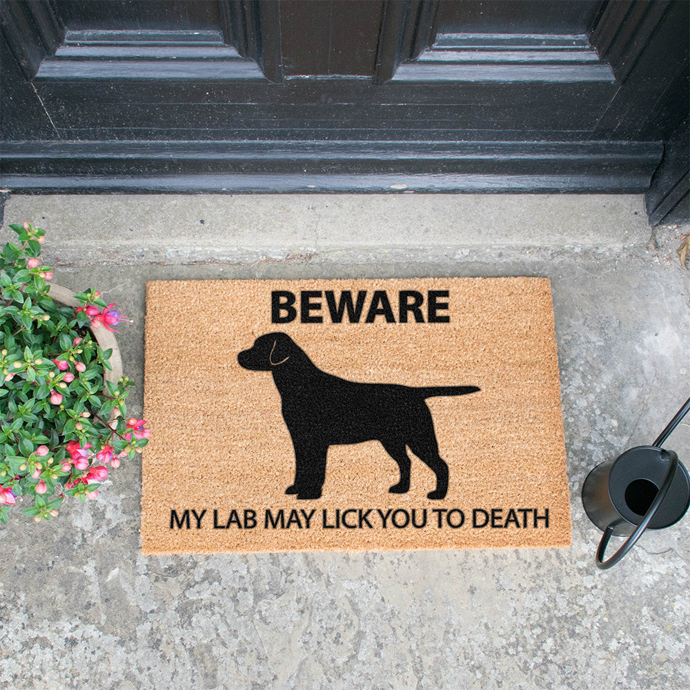 Coir doormat featuring the silhouette of a Black Labrador and the words 'Beware my lab may lick you to death'. The mat is in front of a door with a plant and watering can next to it.