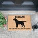 Coir doormat featuring the silhouette of a Black Labrador and the words 'Beware my lab may lick you to death'. The mat is in front of a door with a plant and watering can next to it.